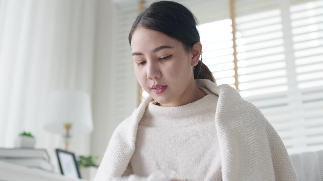 生病的年轻亚洲女人坐在沙发上的毯子下，用纸巾在家里打喷嚏。女性擤鼻涕，咳嗽或在家里用纸巾打喷嚏，患流感。寒热概念视频素材