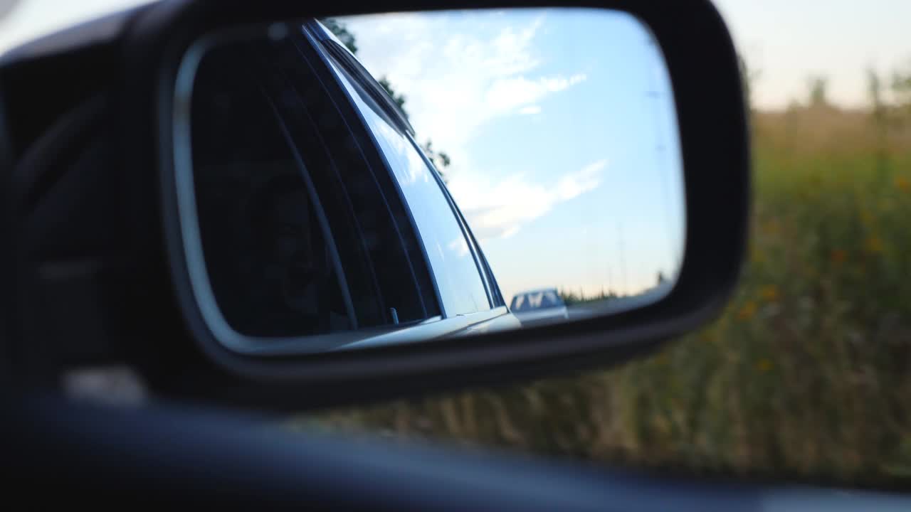 观看道路交通和美丽的天空反射在行驶的汽车的侧镜。夏天，汽车在乡间路上行驶。度假或旅行的概念。特写慢动作视频素材