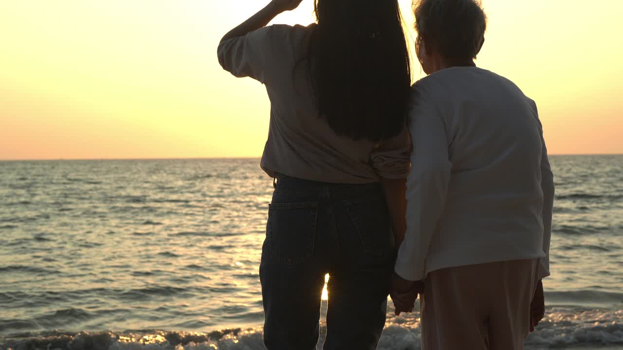 一个女人牵着她妈妈的手在海滩上。他们一起微笑着看日落。视频素材