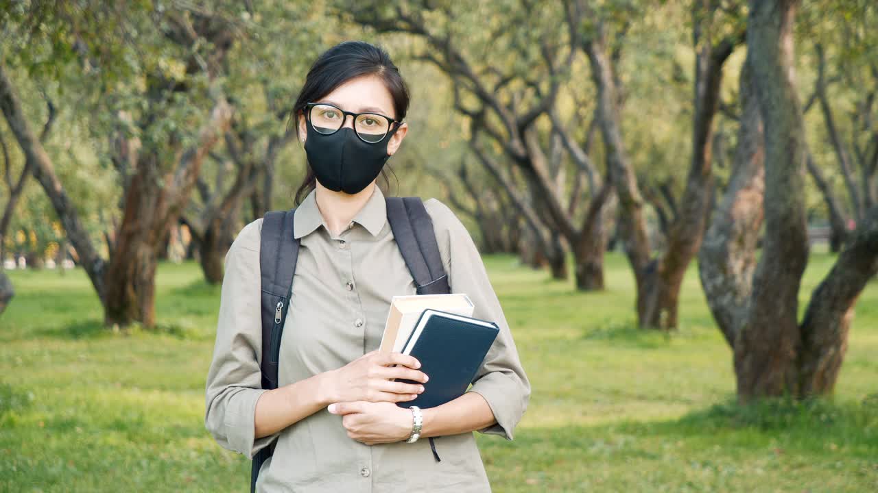 一名年轻女子的肖像，戴着防护面具，拿着一本书，看着户外的相机。大流行期间的教育概念。视频素材