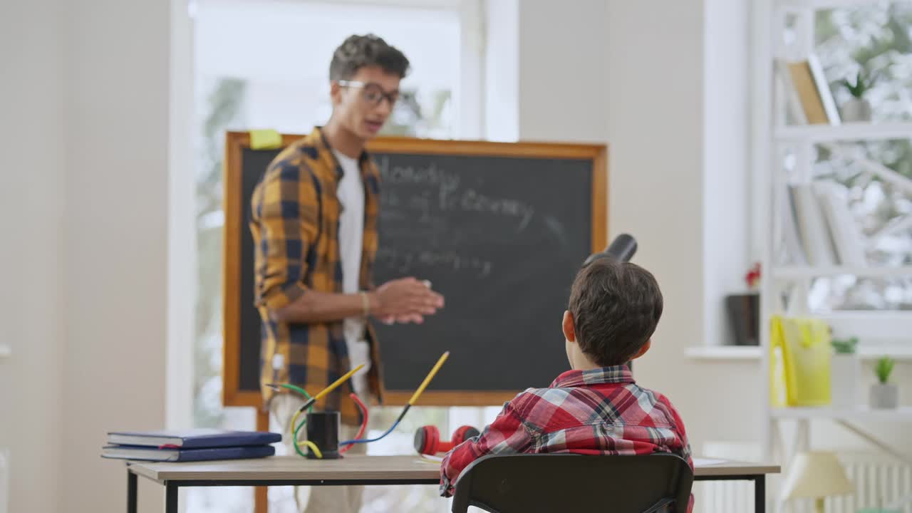 后视图集中的学生坐在桌子上听模糊的年轻老师说话和手势在缓慢的动作。感兴趣的中东男孩在教室学习在学校。情报的概念。视频素材