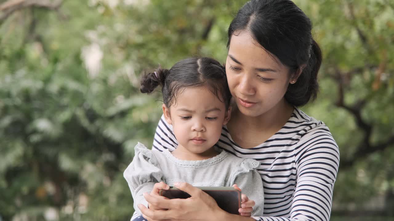 一位亚洲妇女和她的女儿一起在公园玩智能手机。视频素材