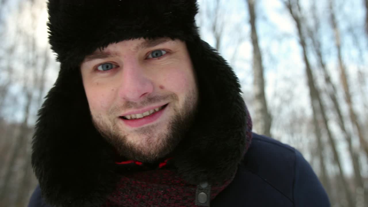 一个滑稽的男人戴着冬天的帽子在下雪的街道上看着相机的肖像视频素材