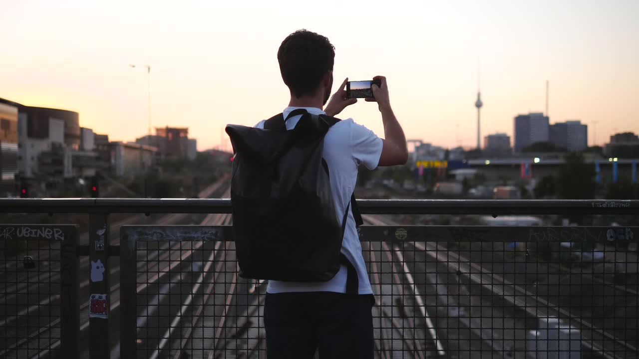 年轻男性游客在日落时站在人行桥上拍摄城市的后视图视频素材