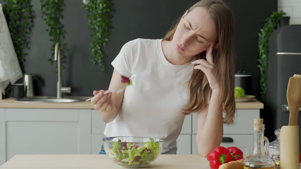 不快乐的女人在厨房的桌子上吃蔬菜沙拉。不高兴的年轻女子吃绿叶生菜视频素材