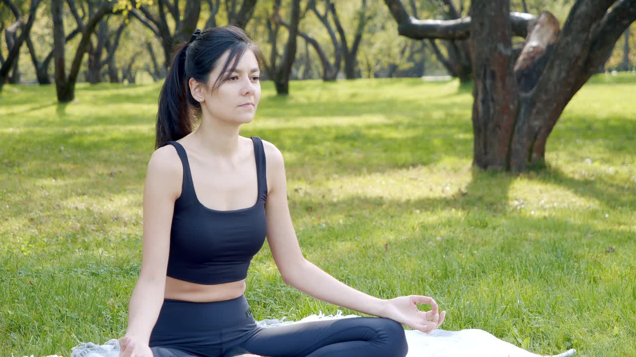 快乐的女人在莲花体位沉思深呼吸，而坐在公园的草地上。瑜伽和和谐的概念。视频素材