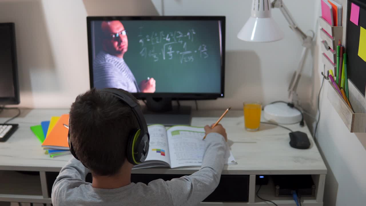 远程学习中，一个男孩戴着耳机坐在家里的桌子旁。理念线上教育、家庭教育、科技、学校视频素材