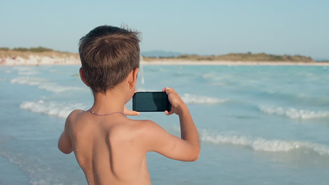 快乐的男孩在海边看屏幕。在海滩上拍摄的少年。视频素材