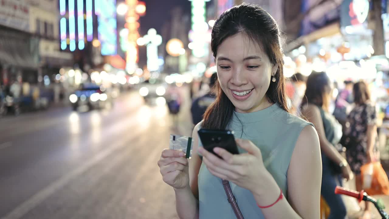 快乐的年轻女子与手机是在网上购买她的信用卡和智能手机在夜间街头市场曼谷路泰国视频素材