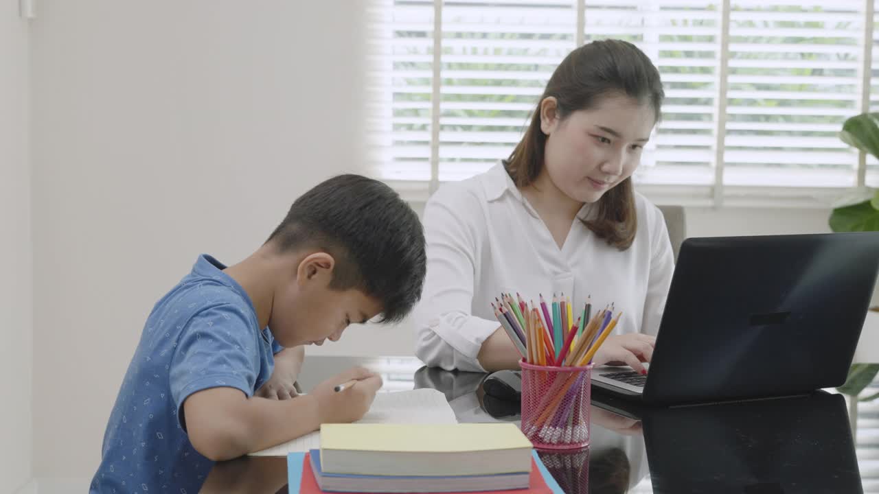 一位亚洲母亲在学校因新冠肺炎疫情而关闭期间，一边在家上学，一边肩并肩工作，教儿子读书视频素材