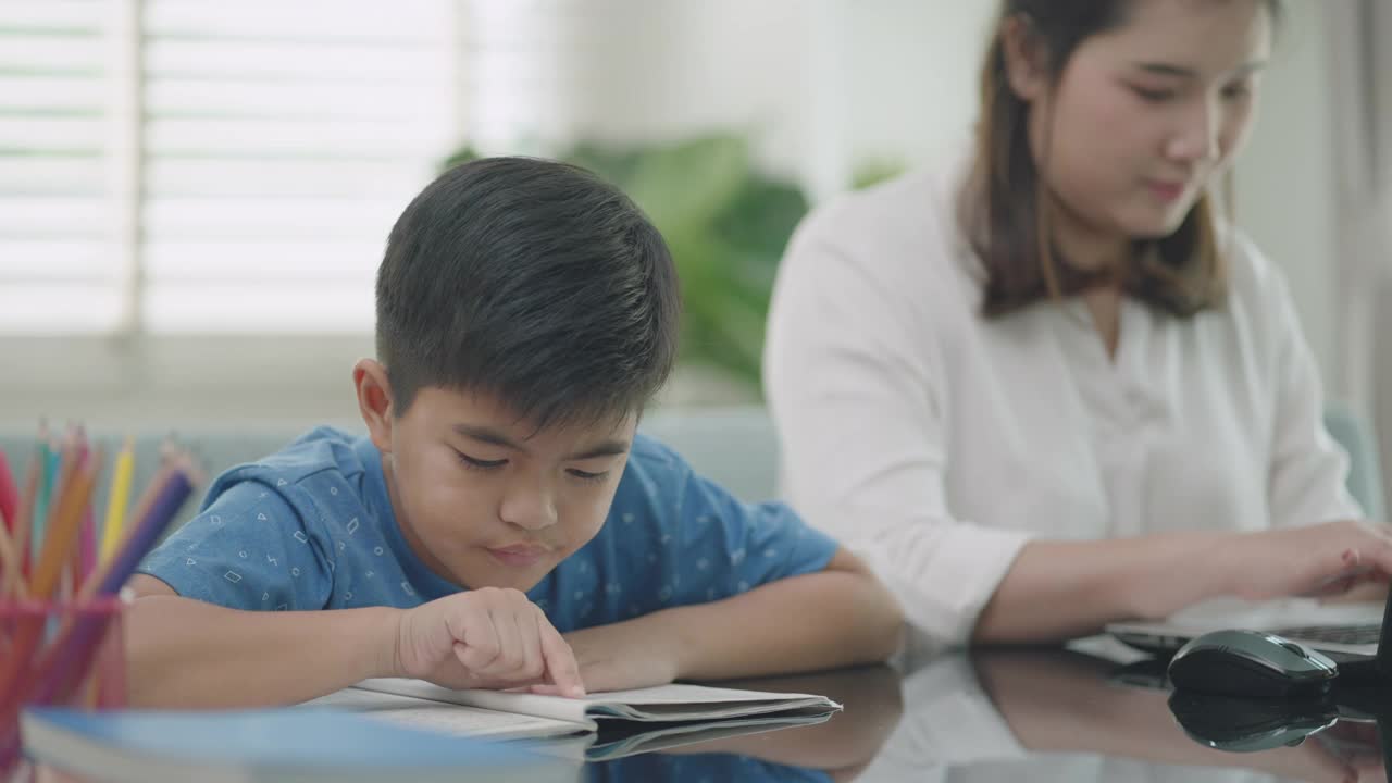 一位亚洲母亲在学校因新冠肺炎疫情而关闭期间，一边在家上学，一边肩并肩工作，教儿子读书视频素材