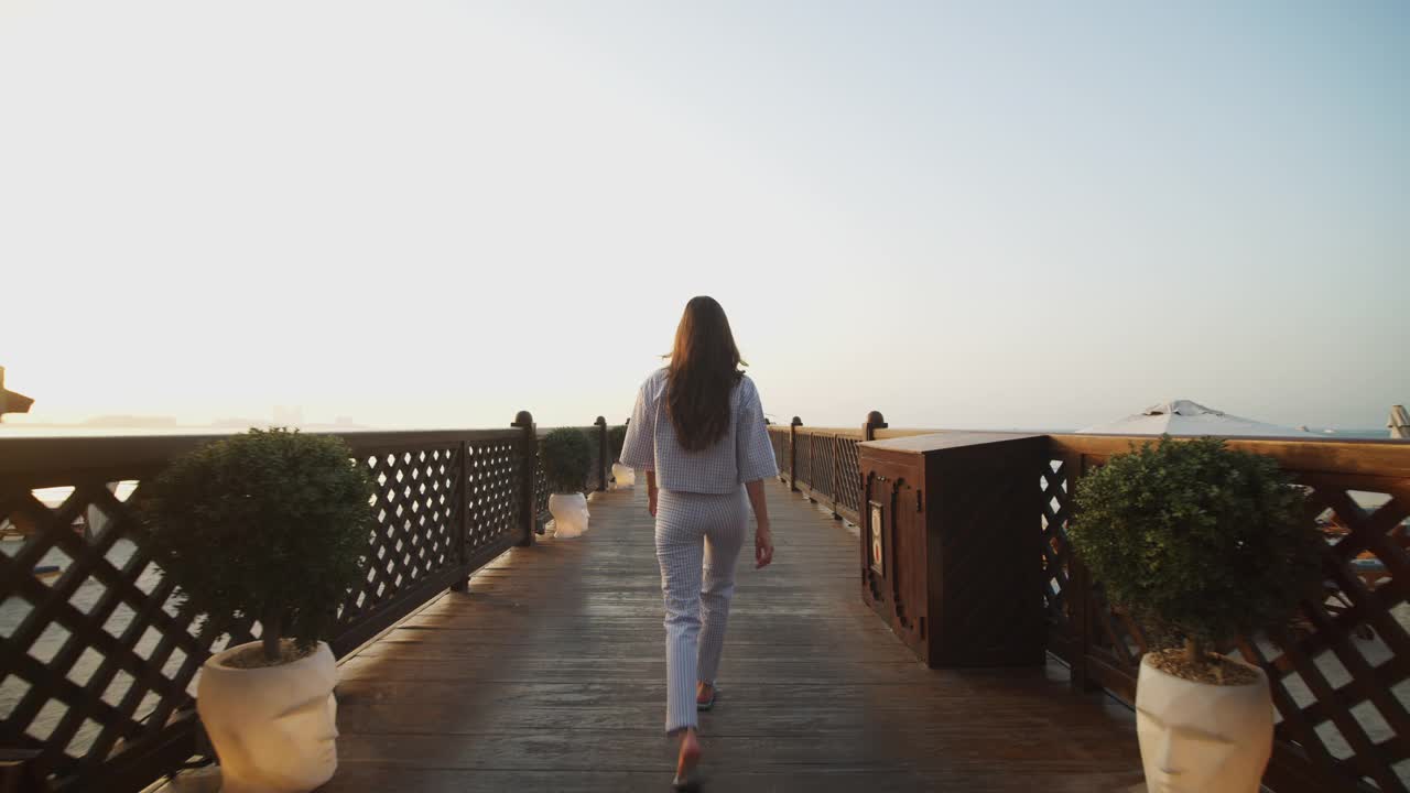 低角度的女人走在木码头在日落。美丽的女游客走在码头上的背影与迪拜的城市景观为背景视频素材