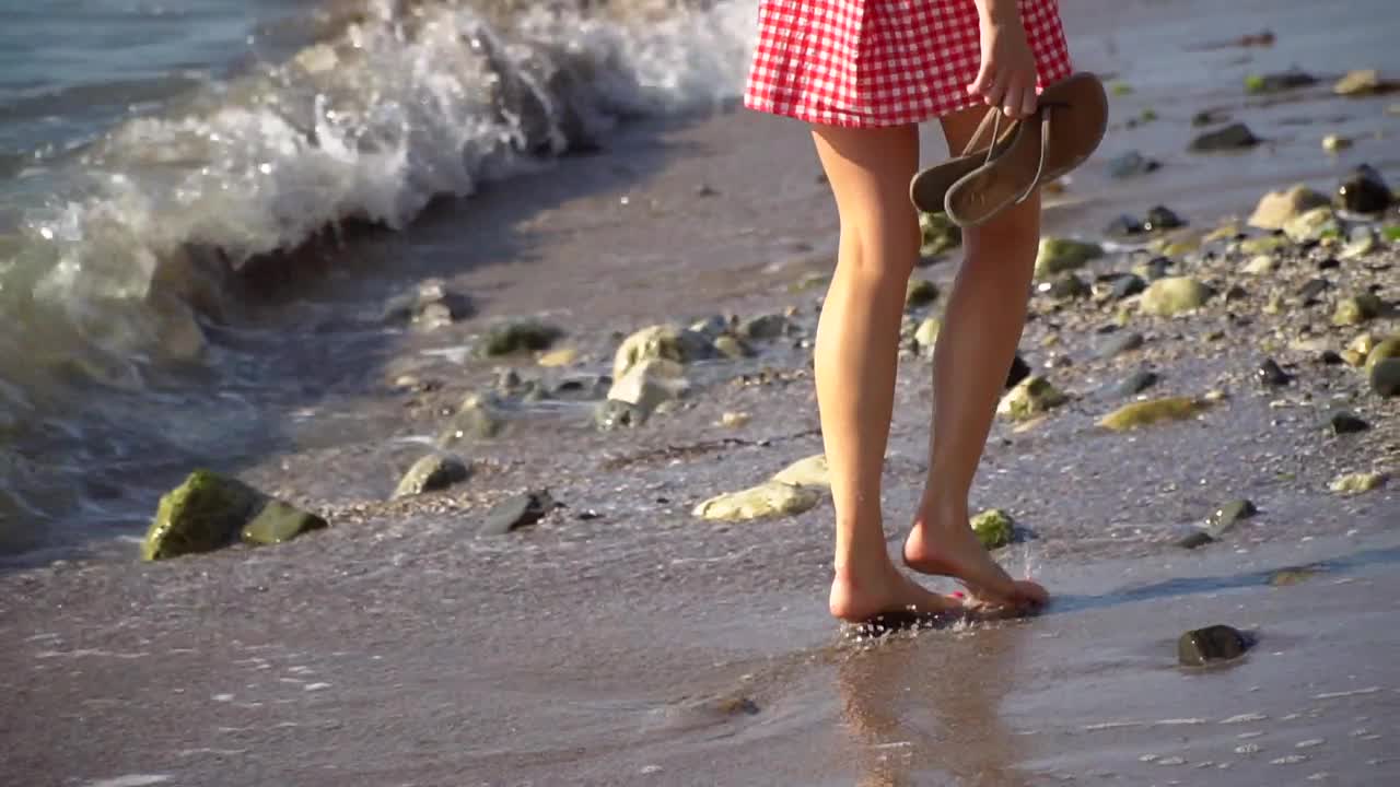 一个女人赤脚在水里沿着海滩奔向大海。女人的脚在水里沿着岸边的石头和火炉走着。那个女孩在水中穿过波浪沿着海滩走。视频素材