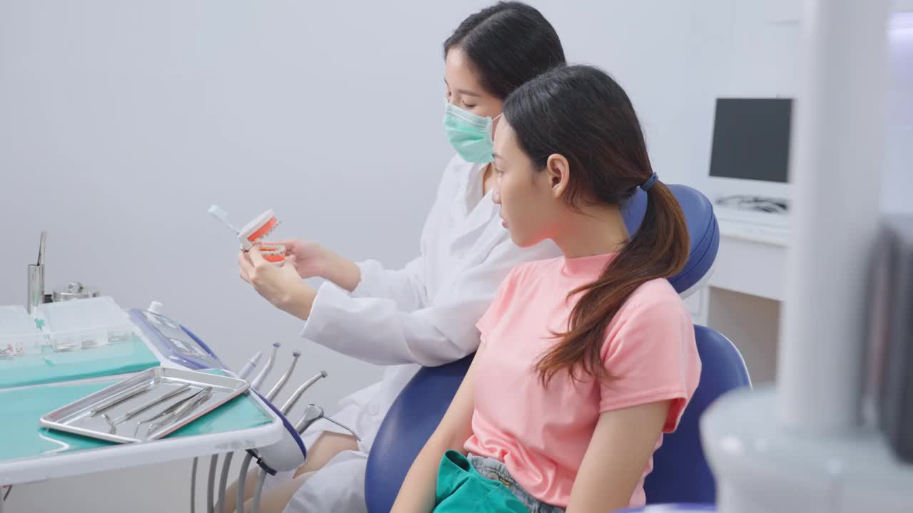 亚洲年轻女孩正在听女牙医讲解口腔护理和牙齿矫正患者的刷牙。牙医在牙科诊所佩戴防护口罩预防COVID-19。视频素材