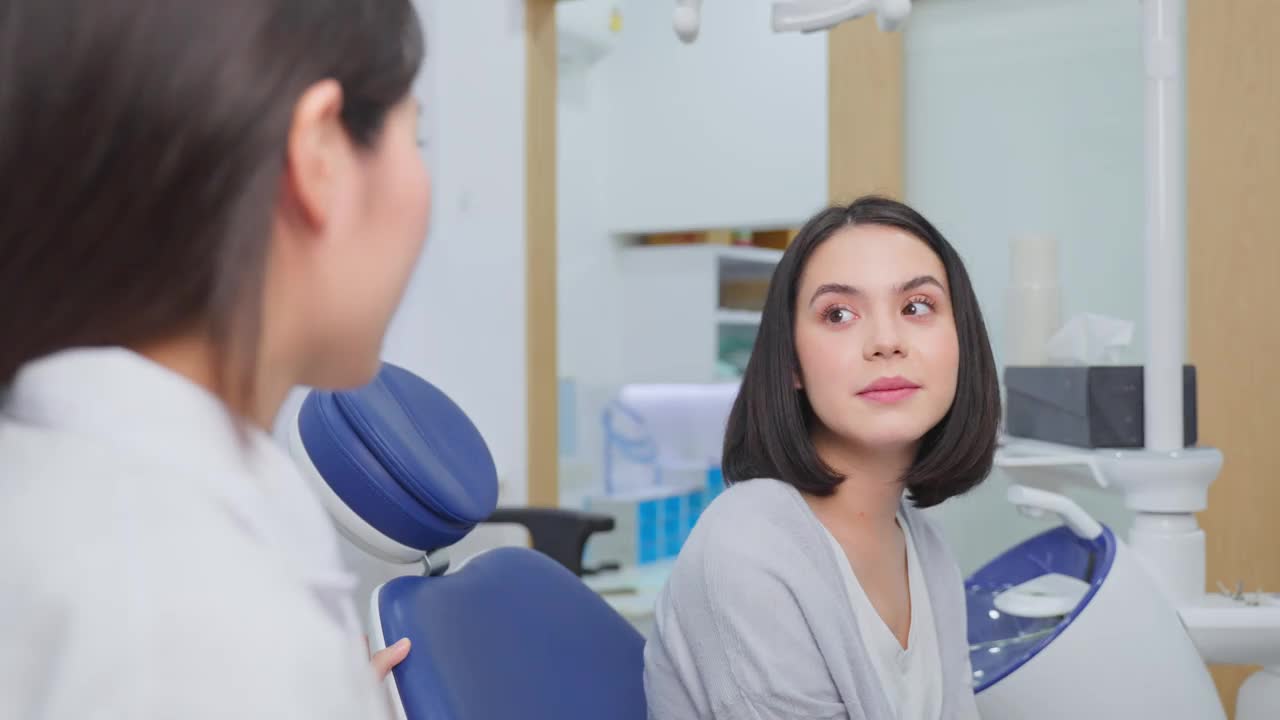 在牙科诊所的牙齿护理计划中，白种美女与亚洲女牙医谈论和咨询龋齿和牙龈疾病。年轻女子很高兴得到专业牙医的照顾视频素材