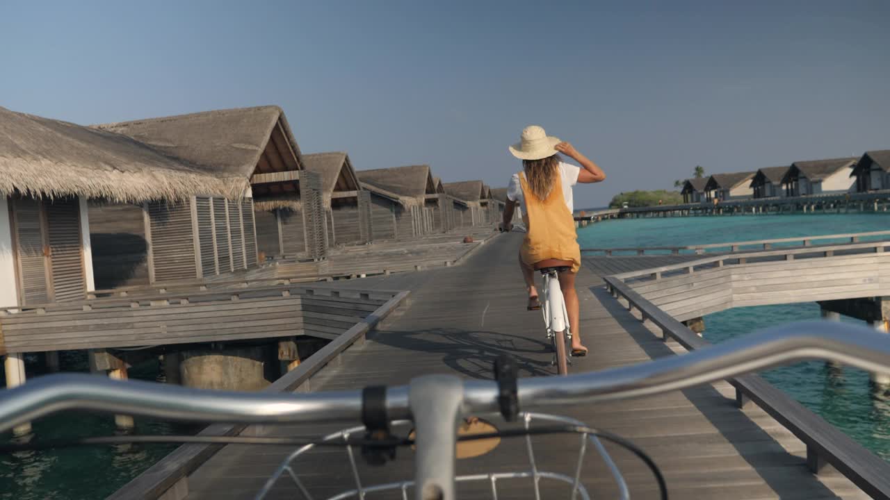 年轻女子骑着自行车在马尔代夫岛的旅游豪华度假村周围。水上平房，女性享受热带度假视频素材