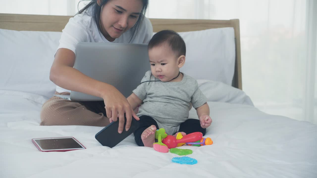 年轻的亚洲妈妈和女儿在卧室的床上与家人一起视频通话，快乐的妈妈和女婴使用智能手机进行教育和互联网在线交流，两人。视频素材