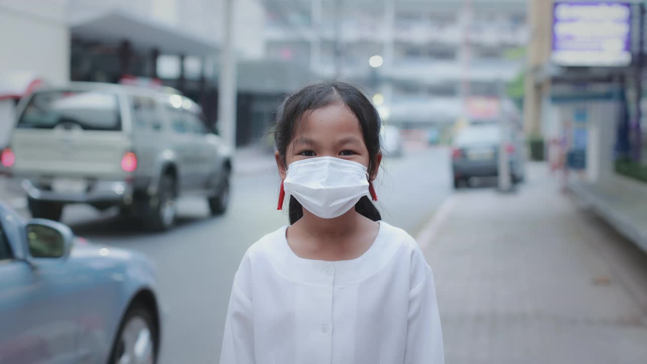 在冠状病毒大流行期间，亚洲女童在口罩后微笑，站在城市的人行道上。视频素材