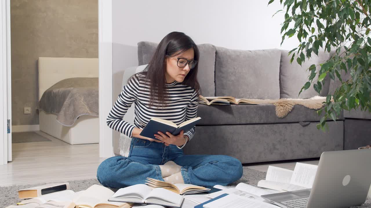 戴眼镜的年轻女学生或记者在许多书籍和教科书中寻找信息，并坐在家里的地板上在笔记本上做笔记。教育的概念视频素材