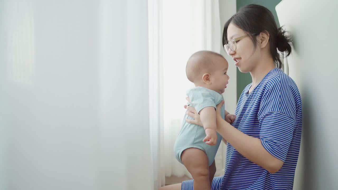 微笑的亚洲年轻母亲抱着她可爱的新生男婴玩。妈妈在家里抱着襁褓中的儿子玩得很开心。家庭、幸福、爱的概念视频素材