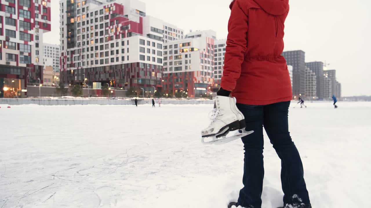 女人手拿着旧的白色冰鞋，看着现代城市的公共溜冰场。冬季户外活动概念。视频素材