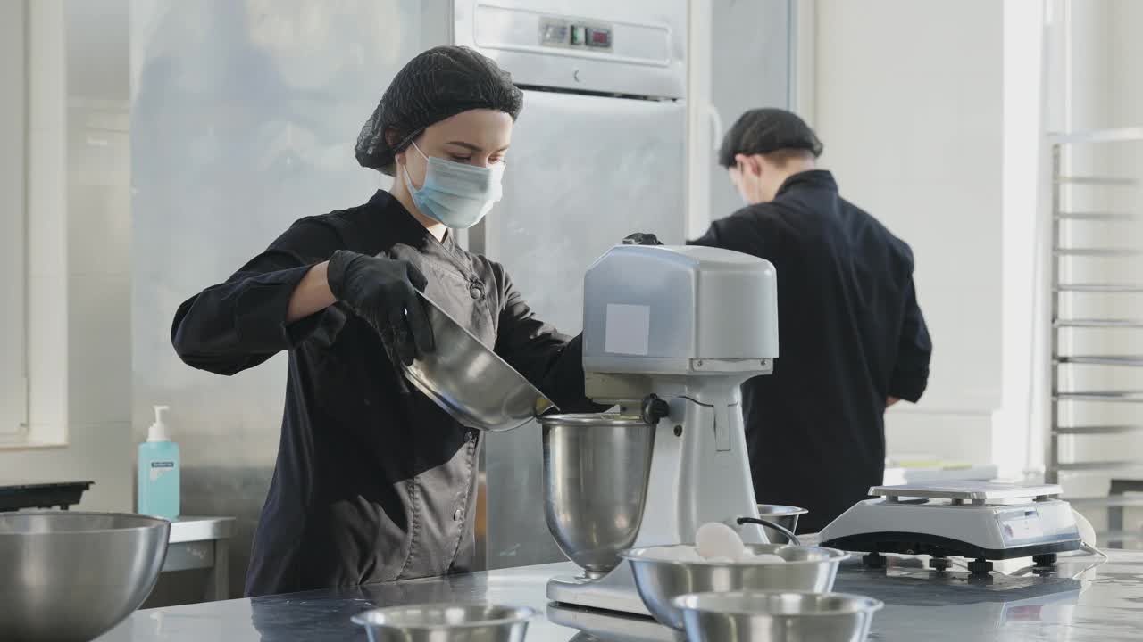 集中的年轻女子在新冠病毒面膜倒配料在自动搅拌机与模糊的男子在商业厨房工作在背景。糖果店工作人员应对冠状病毒大流行。视频素材