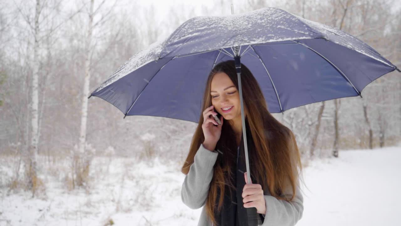 一个年轻漂亮的女人打着伞，打着手机，背景是雪景，正在室外的一个冬天的公园里散步。缓慢的运动。年轻漂亮的女人在冬天的公园里打电话。女性使用智能手机视频素材