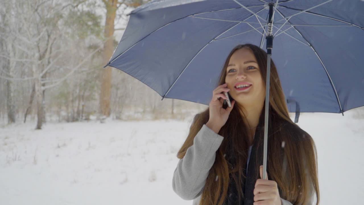 一个年轻漂亮的女人打着伞，打着手机，背景是雪景，正在室外的一个冬天的公园里散步。缓慢的运动。年轻漂亮的女人在冬天的公园里打电话。女性使用智能手机视频素材