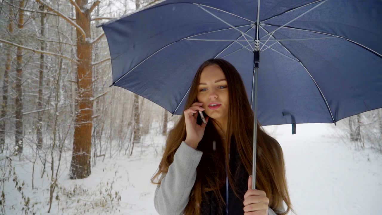 一个年轻漂亮的女人打着伞，打着手机，背景是雪景，正在室外的一个冬天的公园里散步。缓慢的运动。年轻漂亮的女人在冬天的公园里打电话。女性使用智能手机视频素材