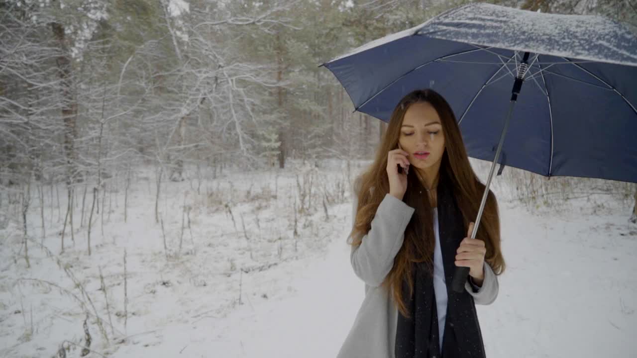 一个年轻漂亮的女人打着伞，打着手机，背景是雪景，正在室外的一个冬天的公园里散步。缓慢的运动。年轻漂亮的女人在冬天的公园里打电话。女性使用智能手机视频素材