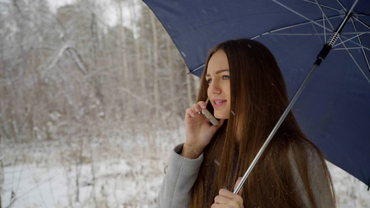 一个年轻漂亮的女人打着伞，打着手机，背景是雪景，正在室外的一个冬天的公园里散步。缓慢的运动。年轻漂亮的女人在冬天的公园里打电话。女性使用智能手机视频素材