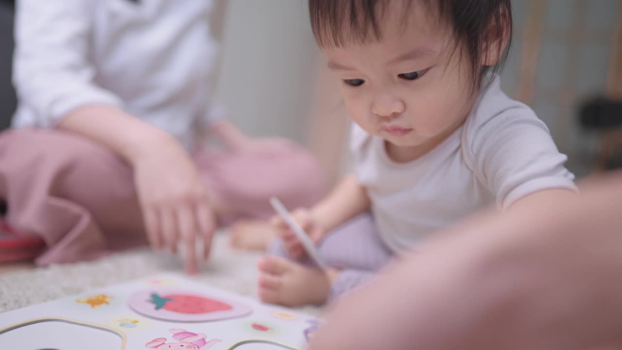 一个顽皮的小女孩在做各种水果形状的纸拼图。年轻的母亲坐在一旁照顾女儿，与家人一起学习活动和快乐时光，一起成长，视频素材