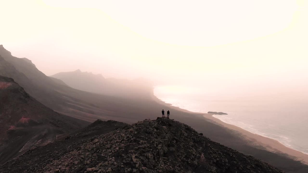 无人机拍摄的两名男子从火山景观的视角俯瞰富埃特文图拉的野生海岸。西班牙。视频素材