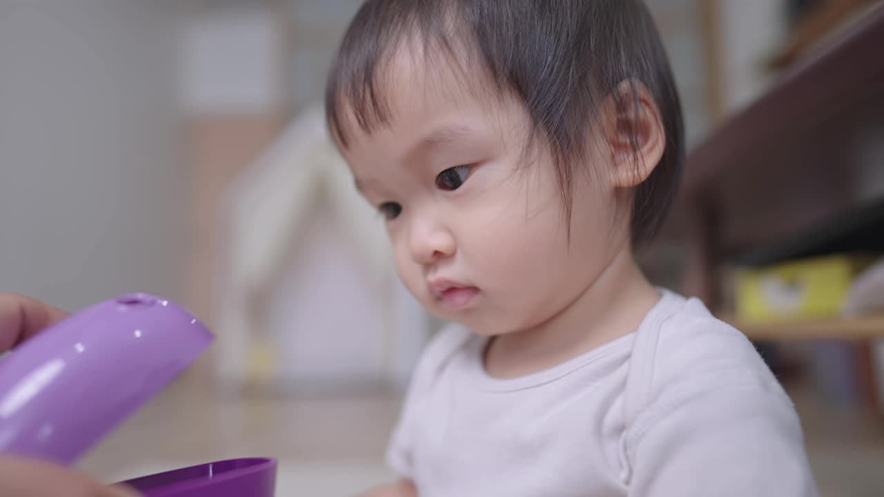 好玩的蹒跚学步的孩子在家里玩的婴儿保姆一天，快乐的小女孩看紫色的玩具和牙齿的微笑，然后愉快地抓住那个玩具，保姆服务在家里视频素材