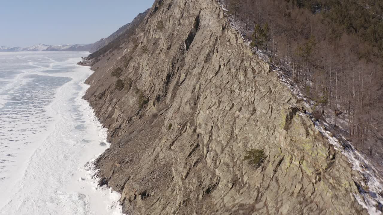 一架无人机在结冰的贝加尔湖岸边的一块长满树木的岩石前飞行。视频素材