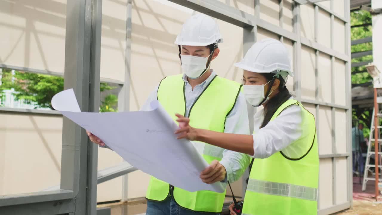 亚洲工人在建筑现场佩戴防护口罩。男和女工程师拿着图纸在施工方讨论项目。视频素材