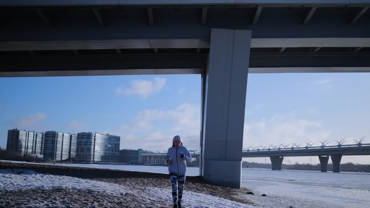 女孩在冰冷的河流旁边的桥下跑完，停止了运动手表上的身体活动分析。视频素材