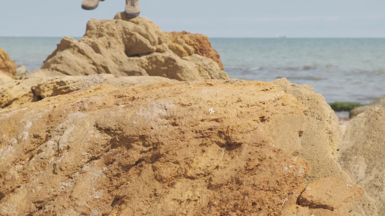 在一个美丽的夏日，年轻的游客穿着靴子偷偷地沿着岩石海岸走。视频素材