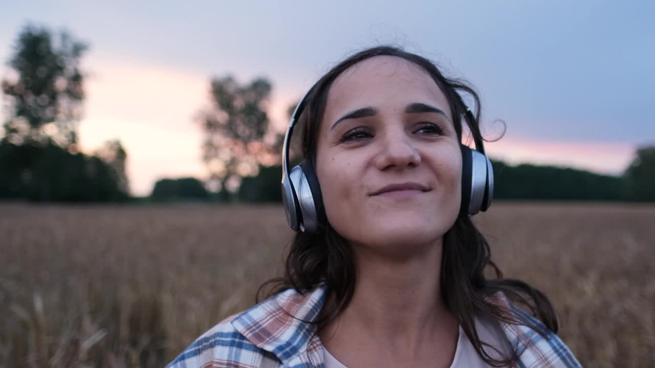 一个穿着格子衬衫的女人戴着耳机听音乐，在日落时分在户外跳舞的慢视频视频素材