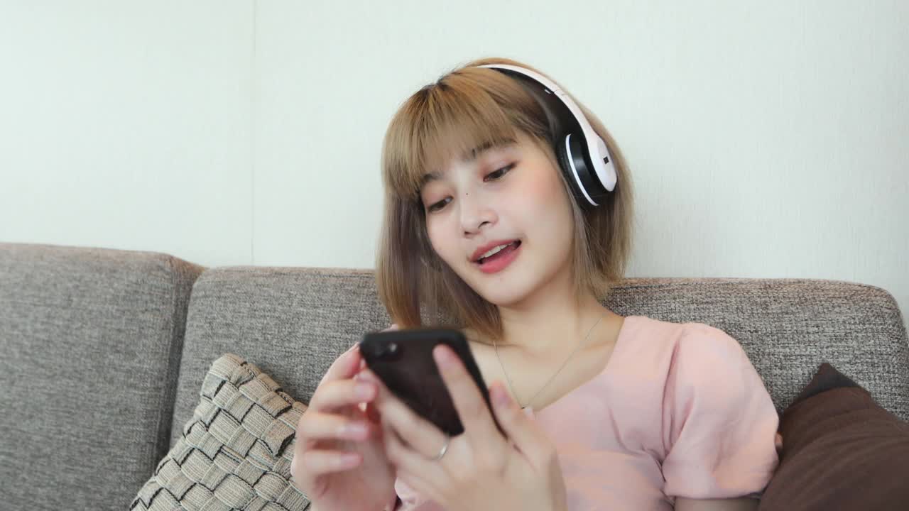 亚洲女性喜欢在家里放松的时候戴着耳机在手机上听音乐视频素材