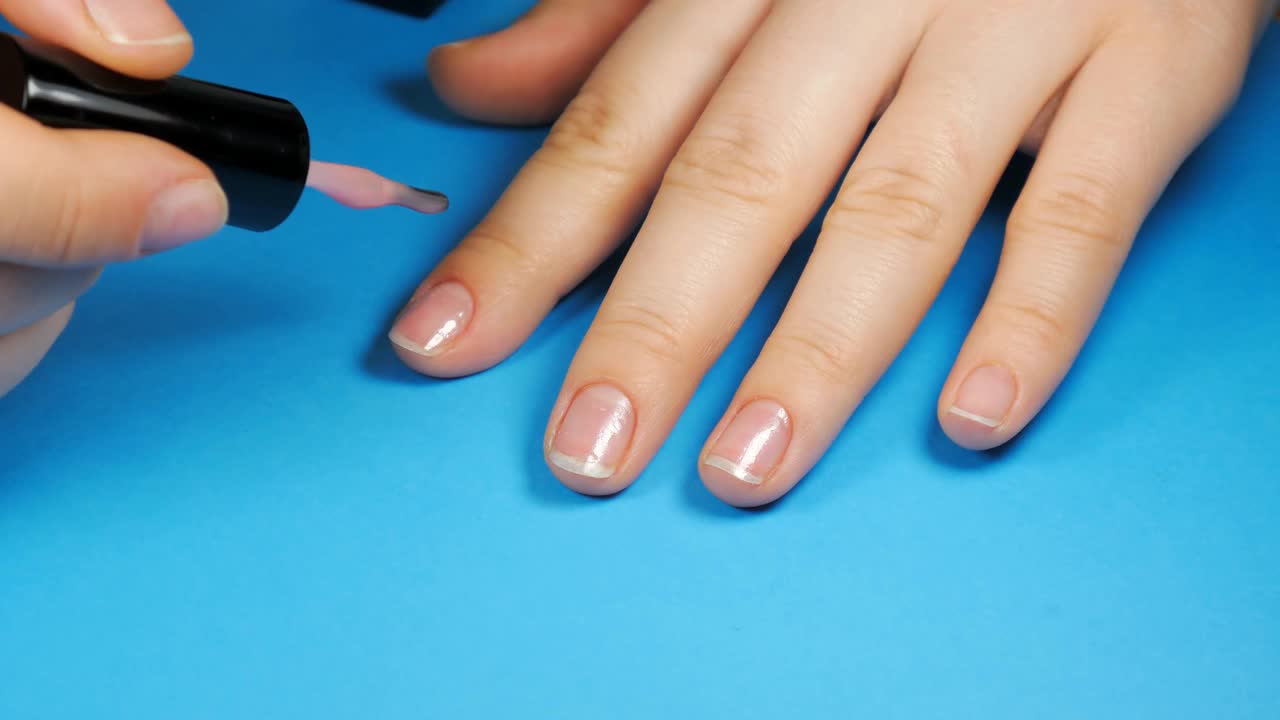 一个小女孩在家里的蓝色背景上涂着浅粉色的指甲油。给自己做个美甲。视频下载