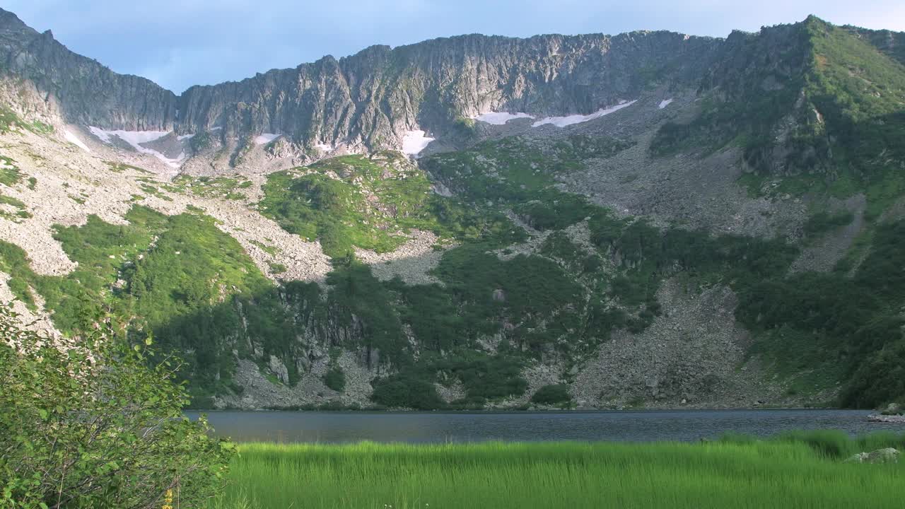 绿色躺在山湖的岸边。风吹皱了草和树叶，使水面泛起涟漪。在蓝天下的地平线山脊上视频下载