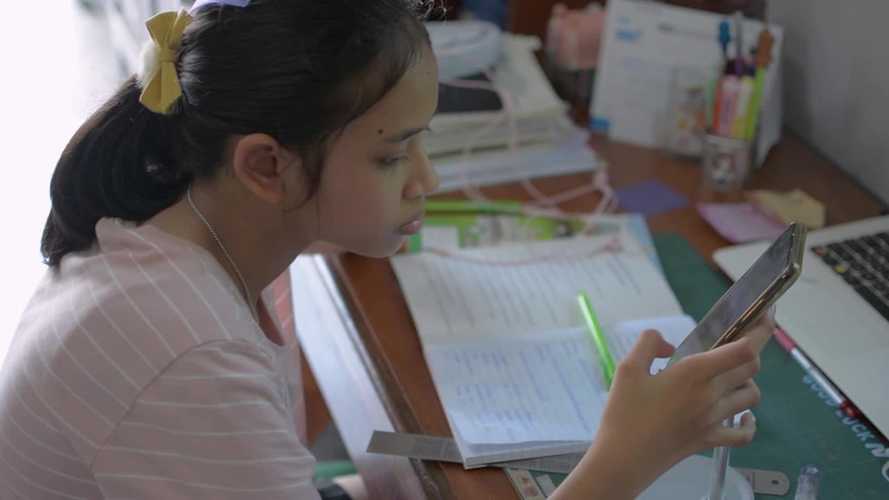 可爱的亚洲女学生坐在桌前用智能手机给远程老师发短信。高中女孩通过手机app学习在线课程。视频素材