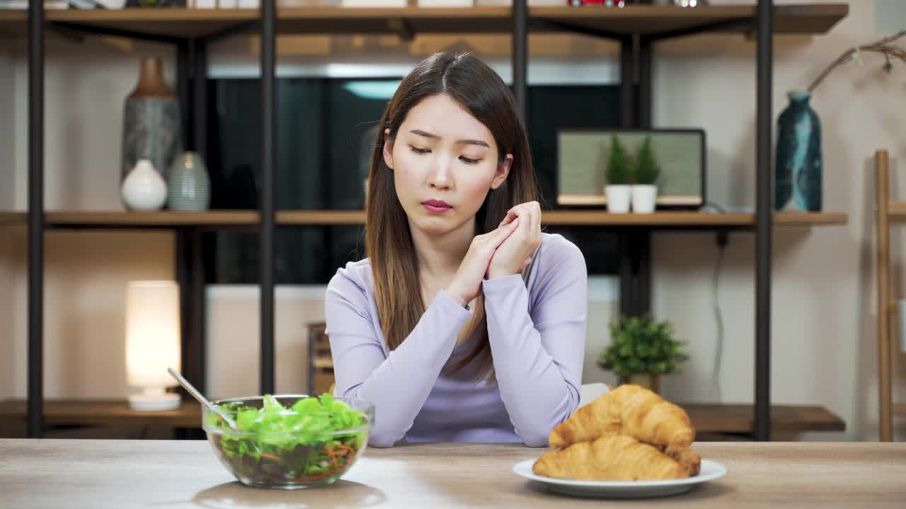 困惑的亚洲妇女犹豫着选择食物。女性选择羊角面包吃满足视频素材