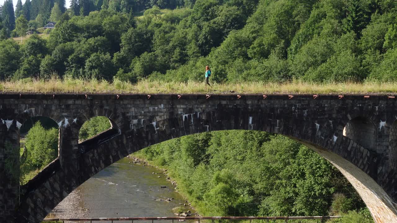 在晴朗的夏日里，一个徒步旅行的女孩穿过山河上古老的石桥视频素材