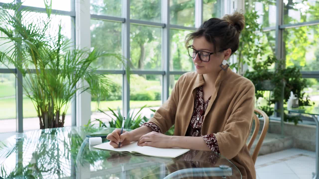 一位严肃美丽的商业女性，一头黑发，戴着现代眼镜，穿着棕色斗篷，戴着耳机，在舒适的浅绿色咖啡馆里用智能手机听音乐视频下载