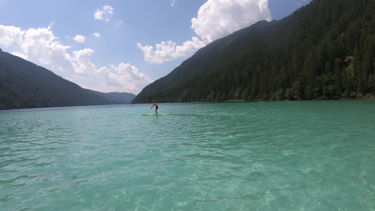 一个阳光明媚的日子，一名男子站在奥地利威森西河上划水。湖的两边都被高山环绕着。山坡上长满了茂密的森林。暑假。水上运动。视频素材