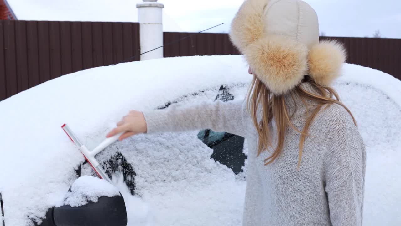 一个戴毛皮帽子、穿毛衣的女孩正在清理车上的积雪。视频下载