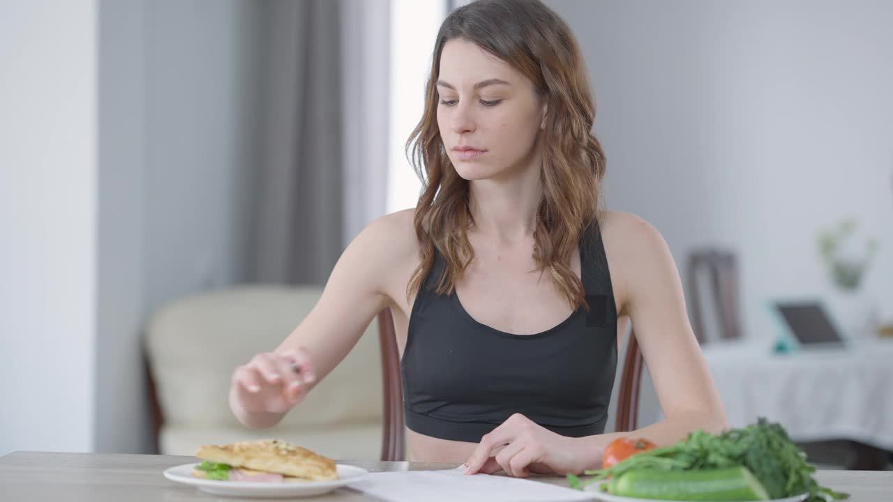 专注苗条的年轻女子写卡路里看着盘子上的汉堡。白种人千禧一代控制体重照顾健康的肖像。自信和自我控制视频素材