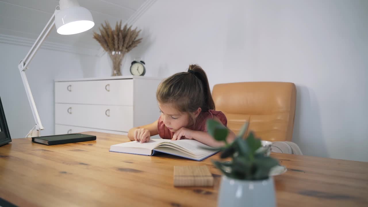 一个女孩正坐在桌子旁看书。这孩子正在学习读书。家庭教育。一个聪明的孩子。读书。孩子们的童话故事。闭关锁国则。摧毁慢动作视频下载